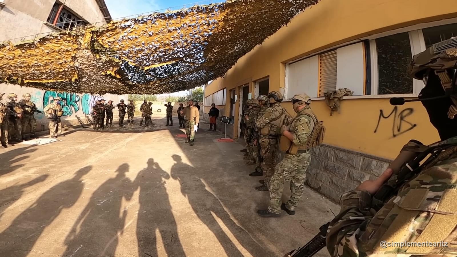 Jogadores equipados e em formatura debaixo de uma rede de camuflagem enquanto o organizador lhes fala antes do jogo