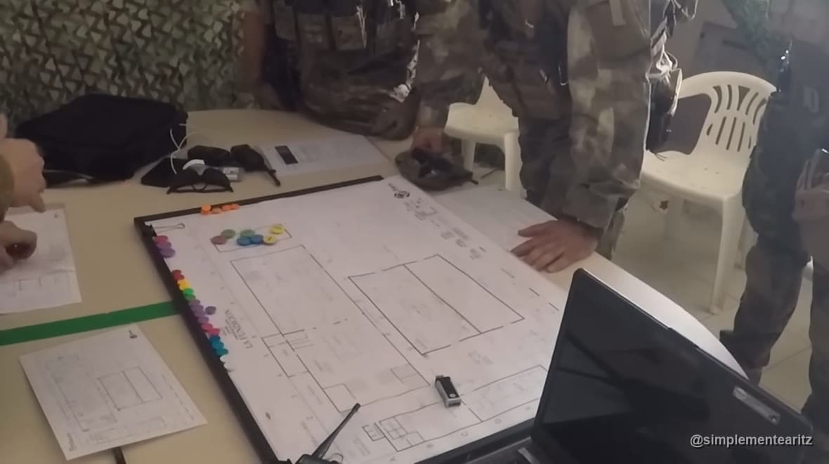 Mesa de briefing com o mapa do campo, fichas de cores e as mãos de vários jogadores