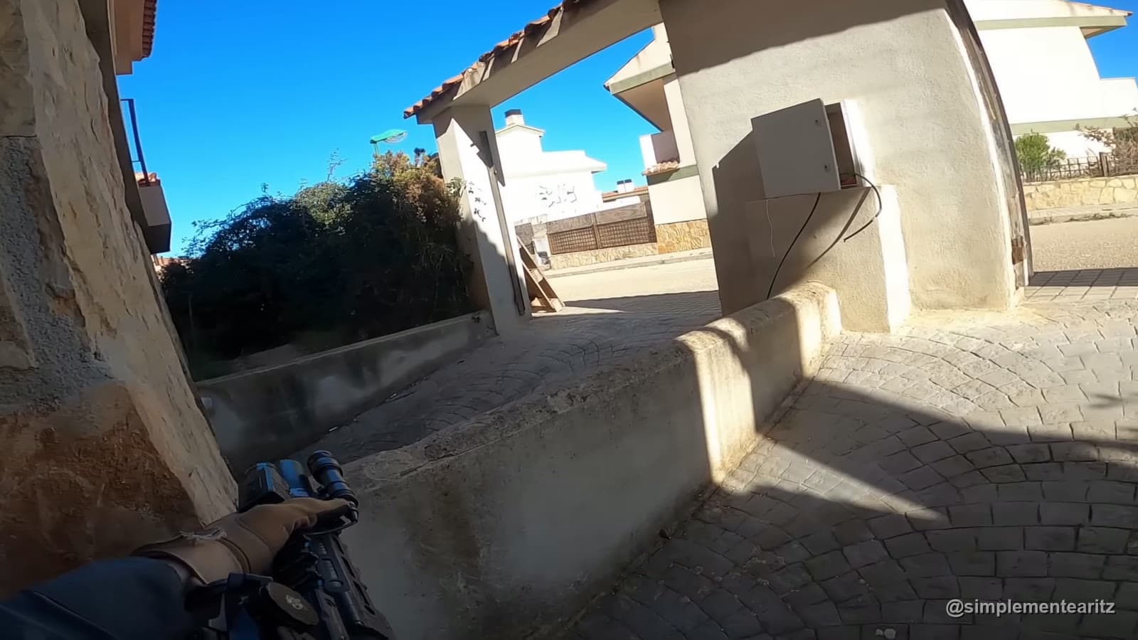 Vista en primera persona de un jugador de airsoft con guantes y réplica, cubierto tras un muro bajo en un campo urbano de casas a pleno sol