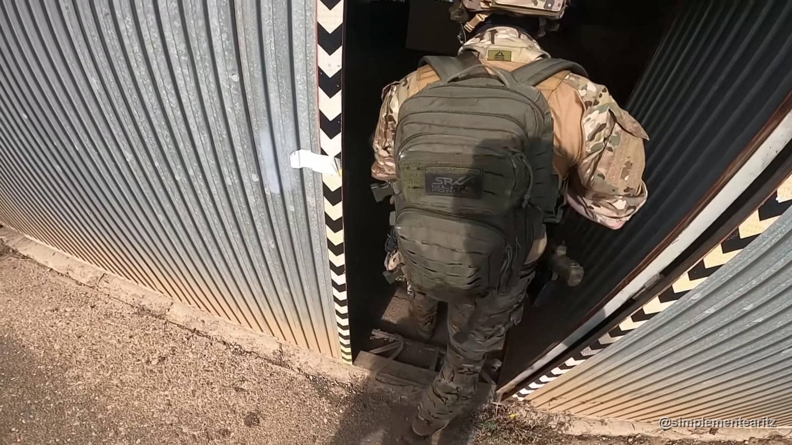 Jogador de airsoft com mochila tática verde e farda multicam a entrar por uma porta estreita de um armazém de chapa ondulada