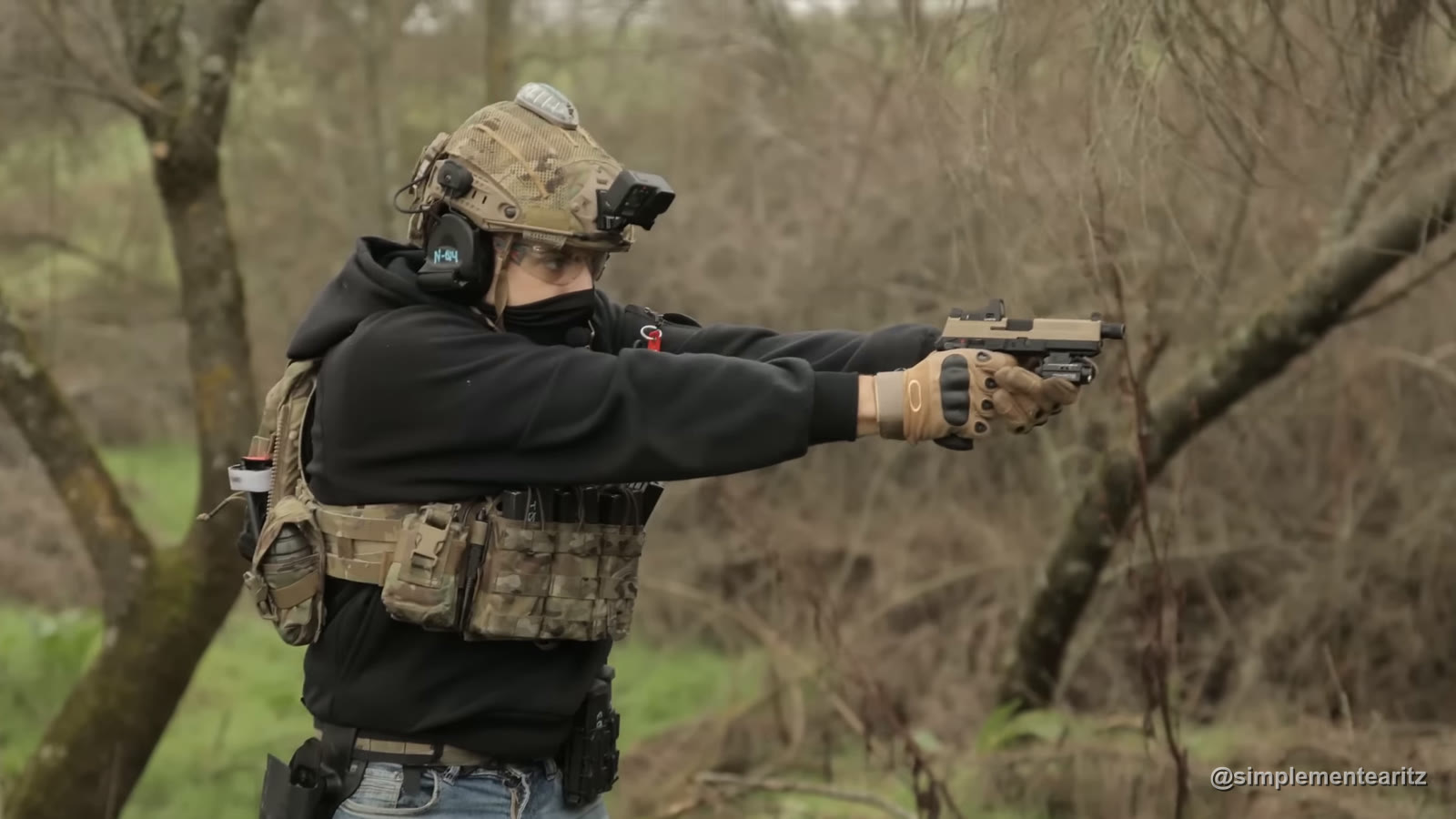 Jugador de airsoft con casco, cascos de comunicación y placa portadora disparando una pistola de gas con visor de punto rojo en un bosque