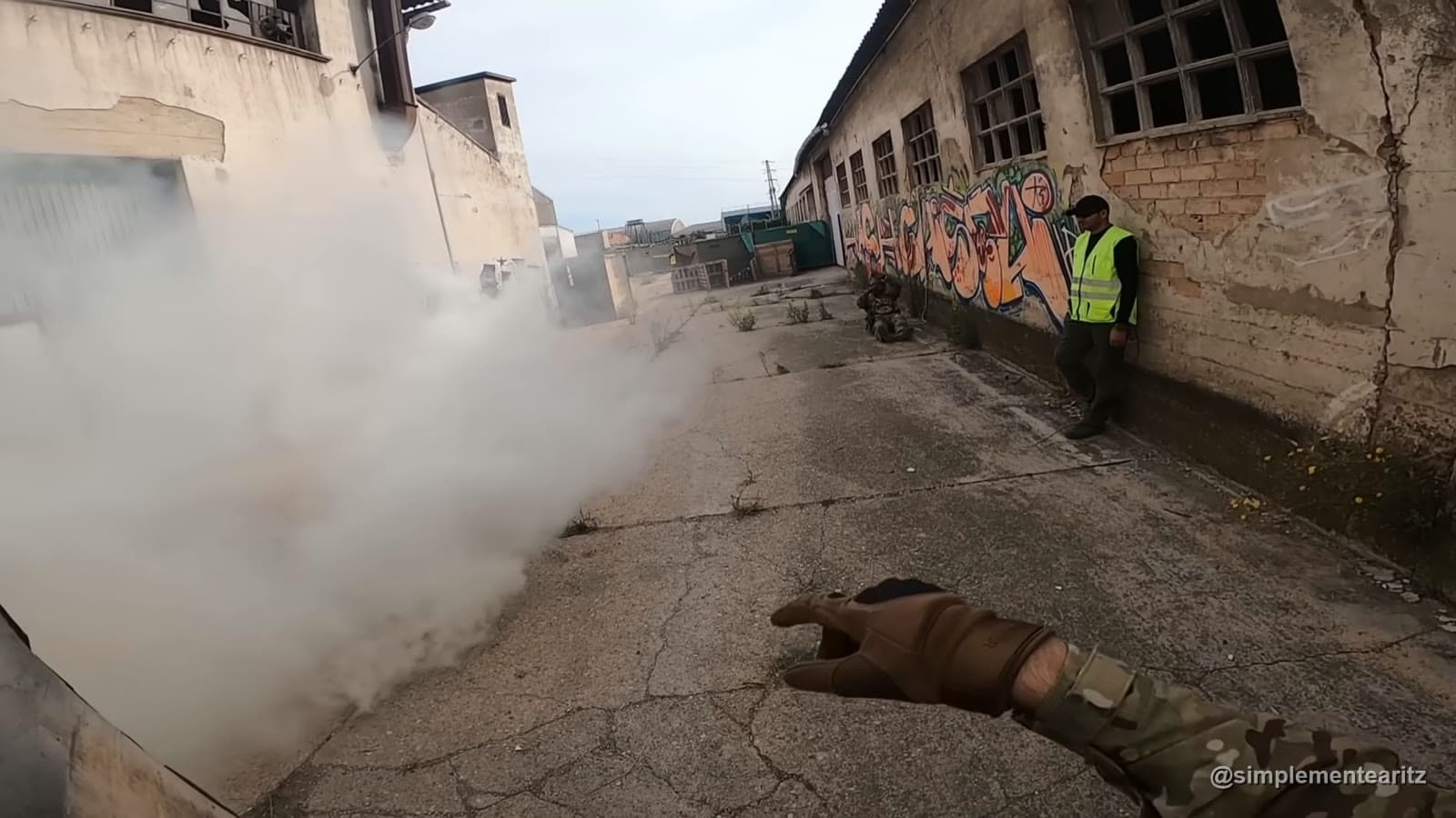 Corredor entre armazéns com fumo, um jogador agachado ao fundo e um árbitro de colete refletor encostado à parede