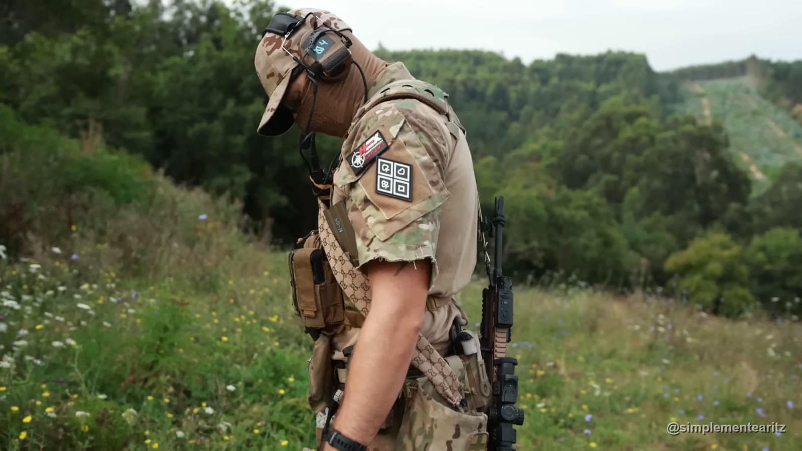 Jugador de airsoft con equipación completa en un campo de bosque, con colinas verdes al fondo