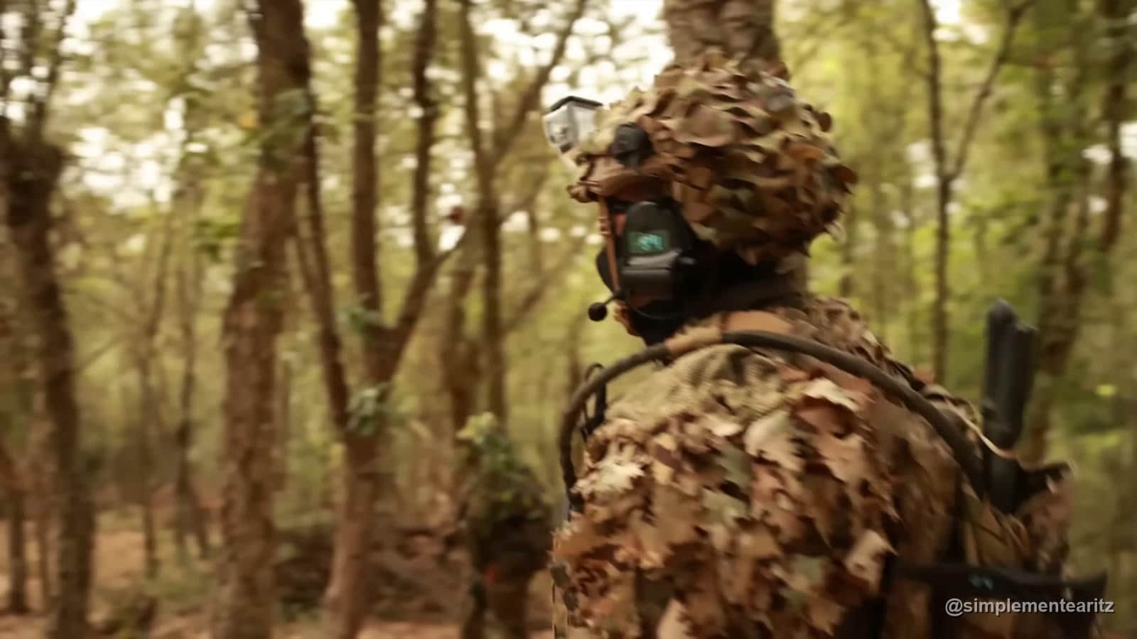 Jugador de airsoft con traje ghillie avanzando entre árboles a contraluz