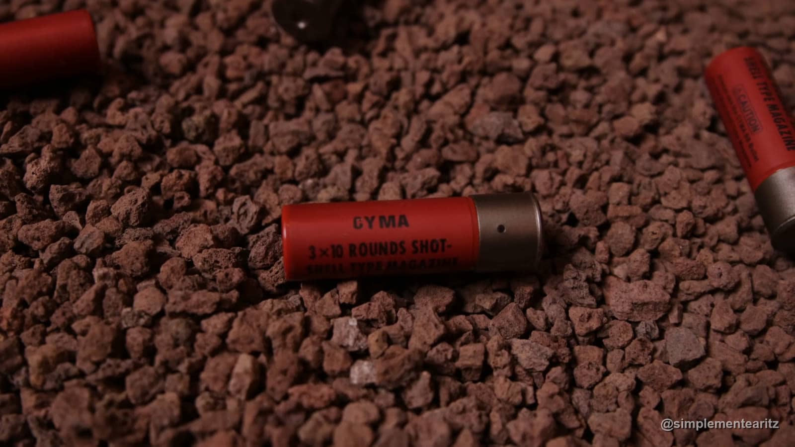 Cartucho rojo de escopeta de airsoft de la marca Cyma, con la inscripción 3×10 rounds shot, sobre grava, junto a otros dos cartuchos