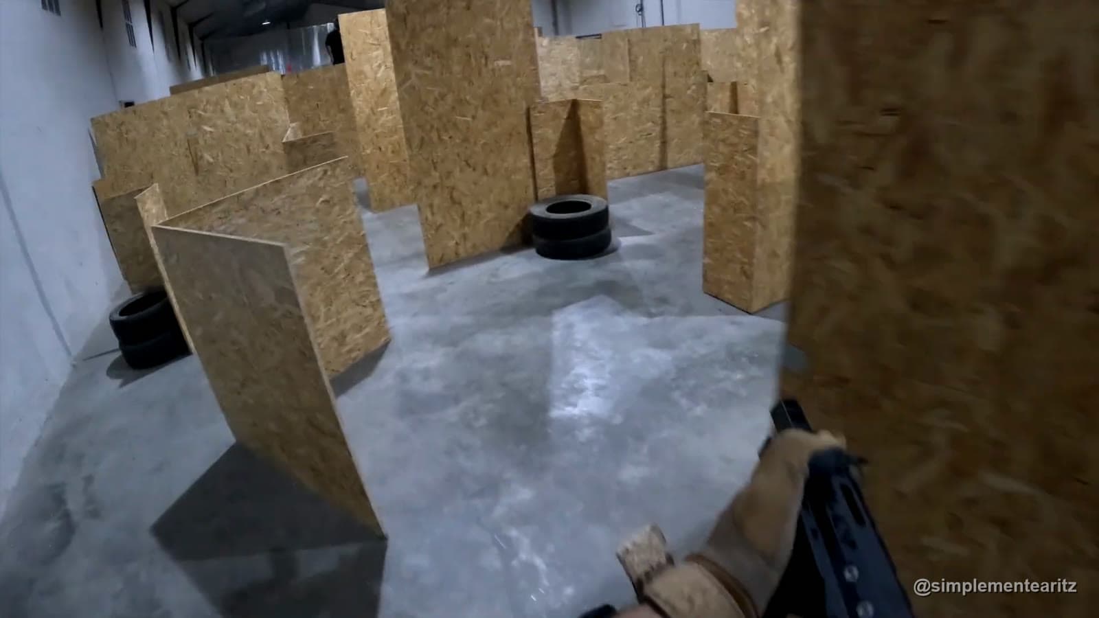 Vista en primera persona de un campo de airsoft de interior en una nave, con tabiques de tablero OSB, neumáticos como parapeto y la réplica asomando por la esquina de un pasillo