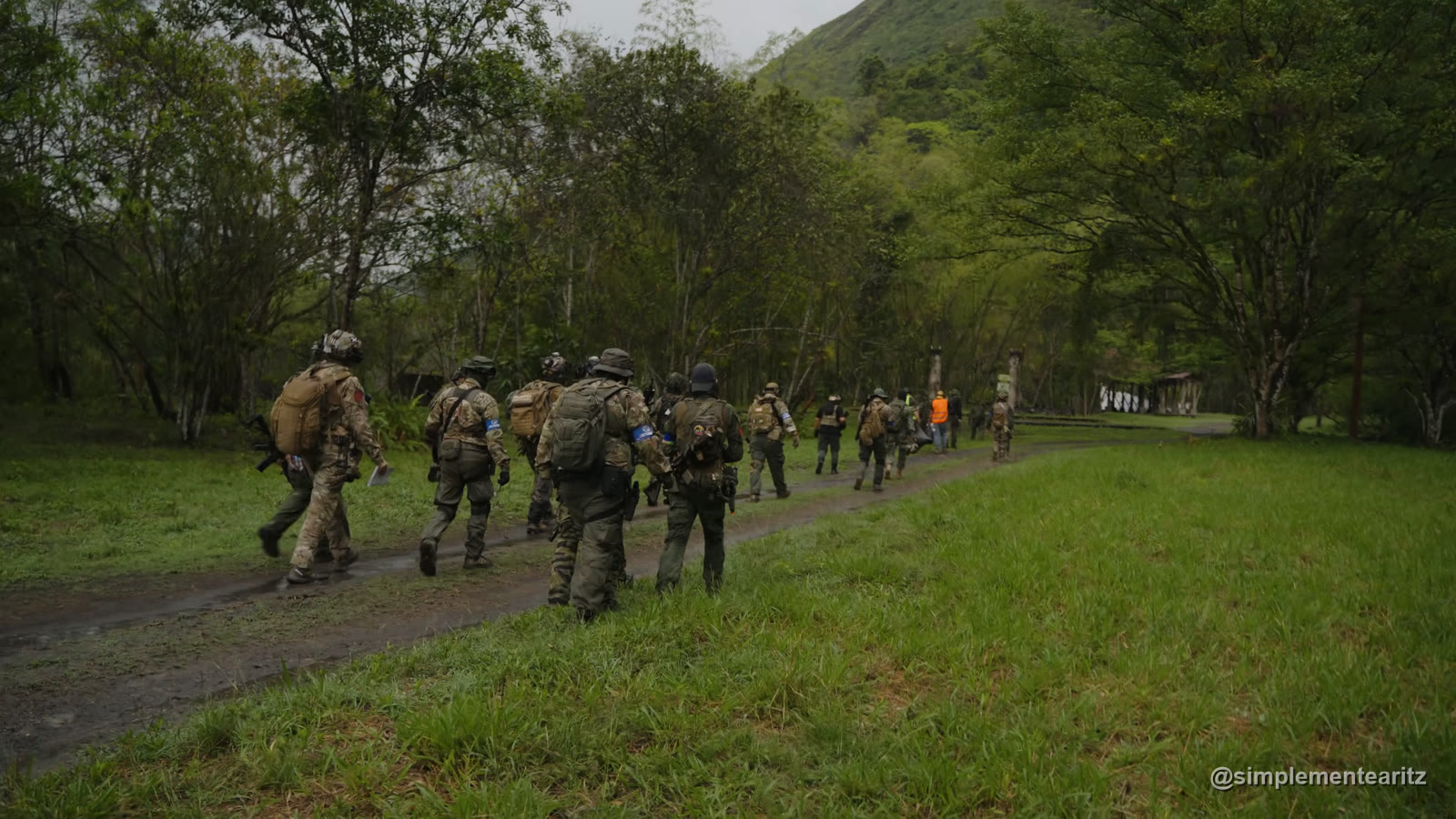Fila de jugadores de airsoft de espaldas, con mochilas y brazaletes azules, caminando por un camino de tierra a lo largo de la explanada de hierba que hacía de campo en la milsim de la selva colombiana, con la montaña al fondo y las lonas blancas de la organización al final