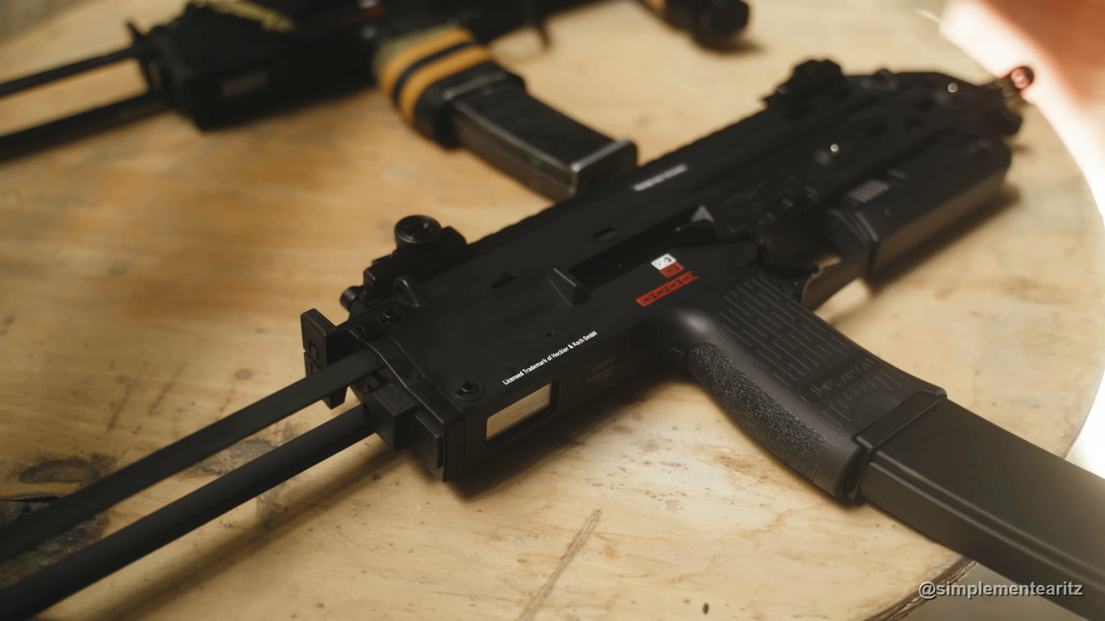 La MP7 de gas de VFC, que vende Umarex con la licencia de Heckler & Koch, negra, sobre una mesa de madera, con «Licensed Trademark of Heckler & Koch GmbH» grabado en el cuerpo; detrás, desenfocada, la de Tokyo Marui con gomas amarillas en la empuñadura