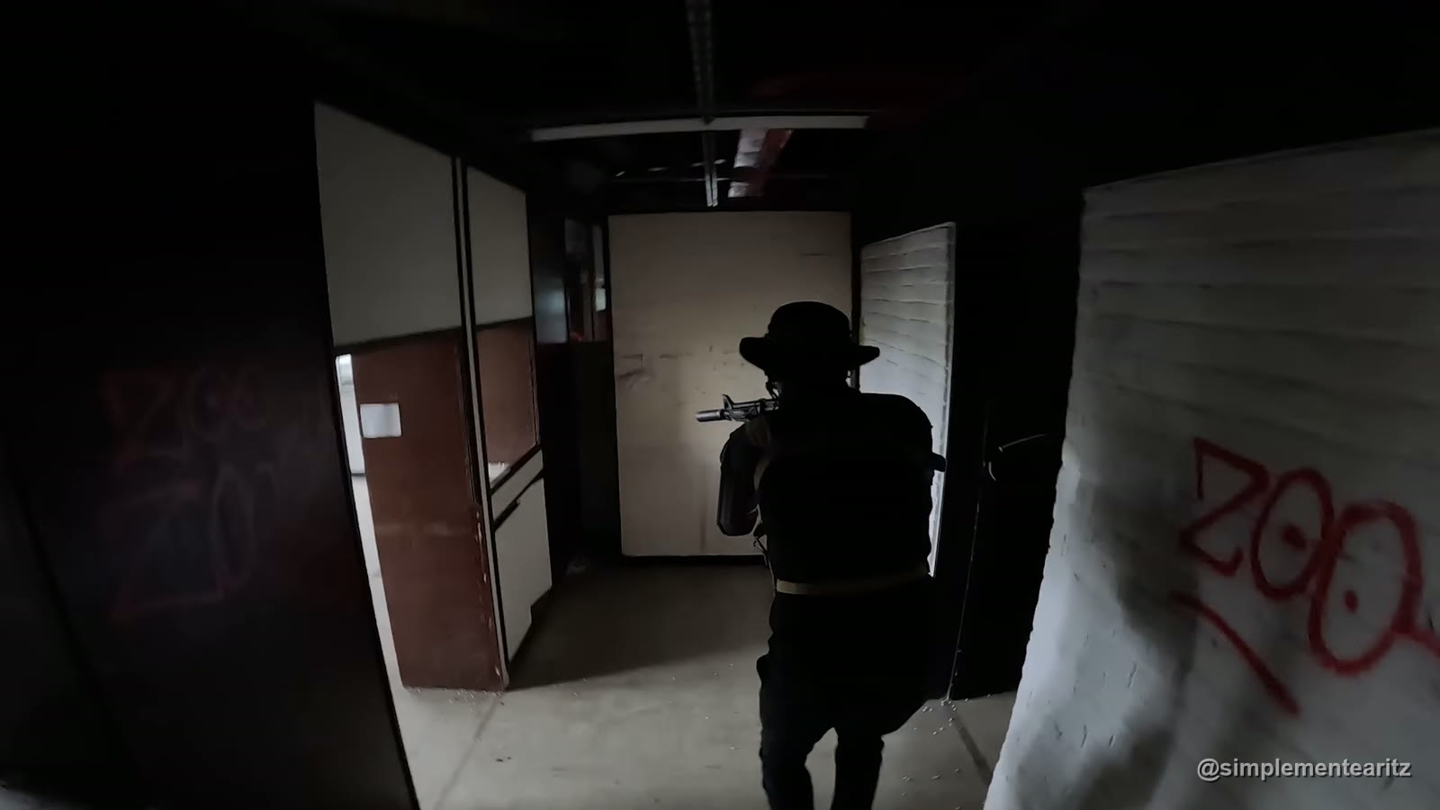 Partida de airsoft en una universidad abandonada de Bélgica, en primera persona: a pocos metros, un compañero con sombrero de ala y el fusil al hombro avanza de espaldas por un pasillo oscuro, entre paredes con pintadas rojas