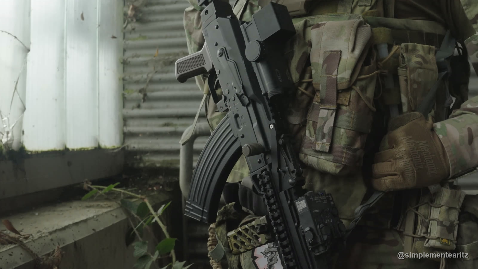 AKM de gas de Tokyo Marui con raíles en el guardamanos y cargador curvo, colgada al pecho sobre un chaleco y un uniforme de camuflaje, junto a una pared de chapa