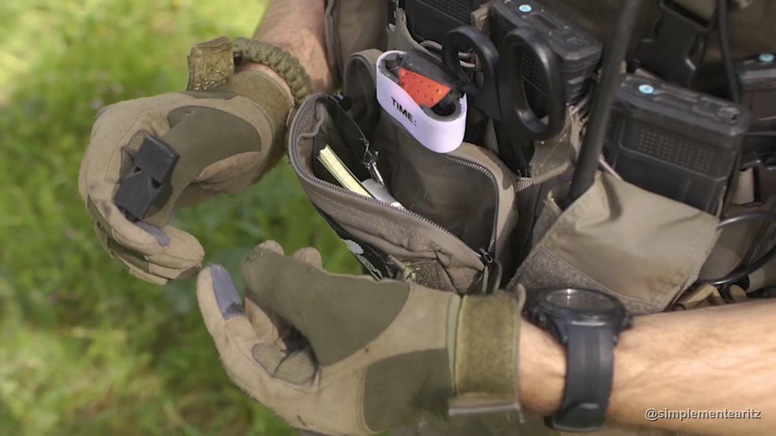 Bolsa de administración abierta en un chest rig verde oliva, con la libreta dentro, uno de los torniquetes con la etiqueta TIME, unas tijeras cortarropa y los cargadores detrás, mientras una mano con guante enseña el silbato