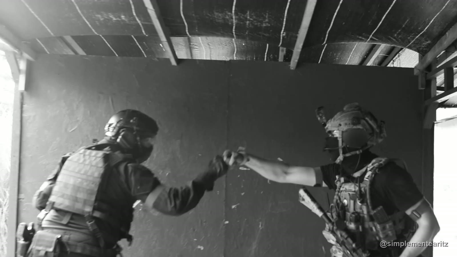 Dos jugadores de airsoft con casco y portaplacas chocan los puños bajo el techo de una caseta en Soldados de Juguete, en blanco y negro