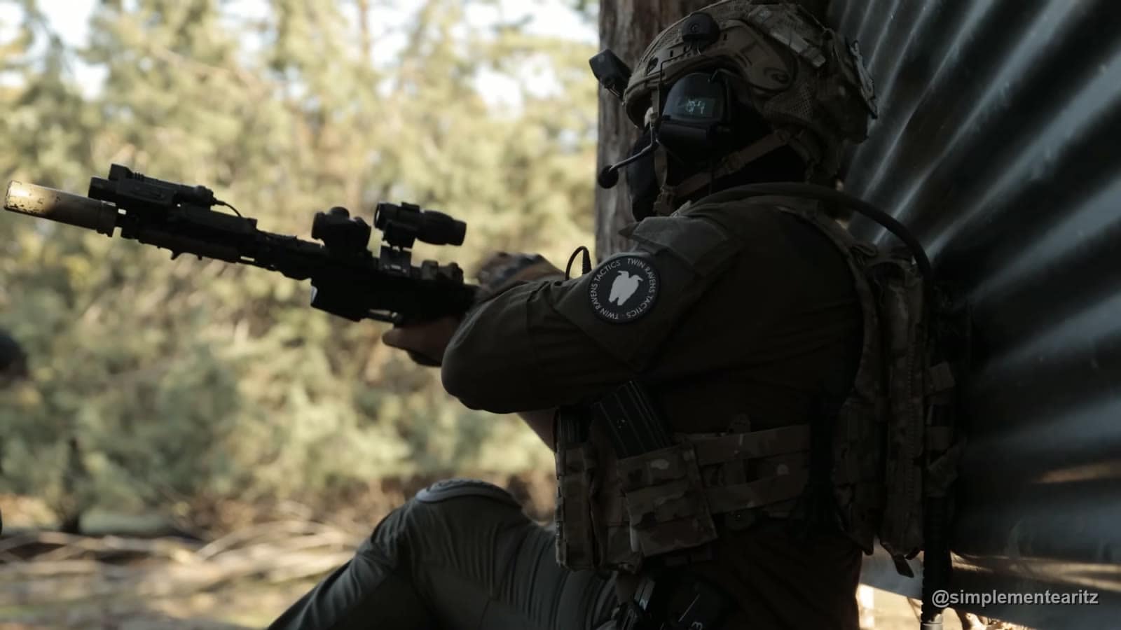 Jugador de airsoft con casco y cascos de comunicación apuntando un fusil tipo M4 con silenciador, visor de punto rojo y magnificador, apoyado en una chapa ondulada