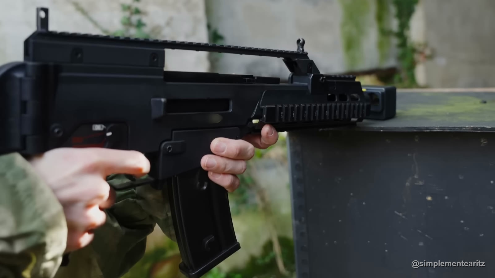 Réplica de airsoft G36 de gas sujeta con las dos manos junto a una caja, con un cronógrafo puesto en la boca del cañón para medir su potencia