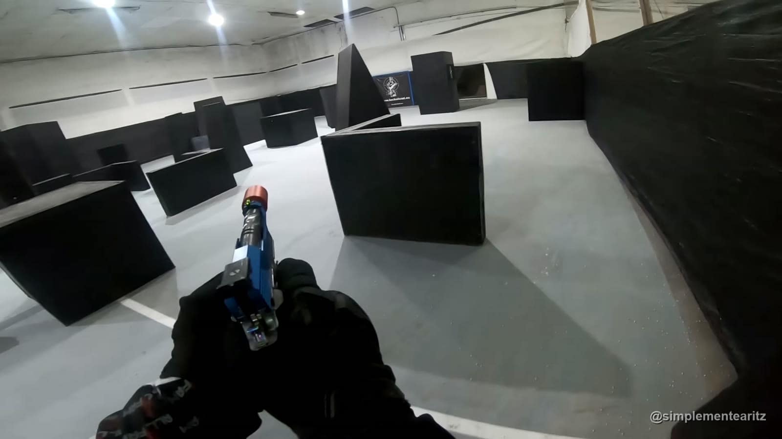 Pistola Hi-Capa azul con unidad trazadora roja en la boca del cañón, vista en primera persona en un campo de speedsoft de interior con obstáculos negros
