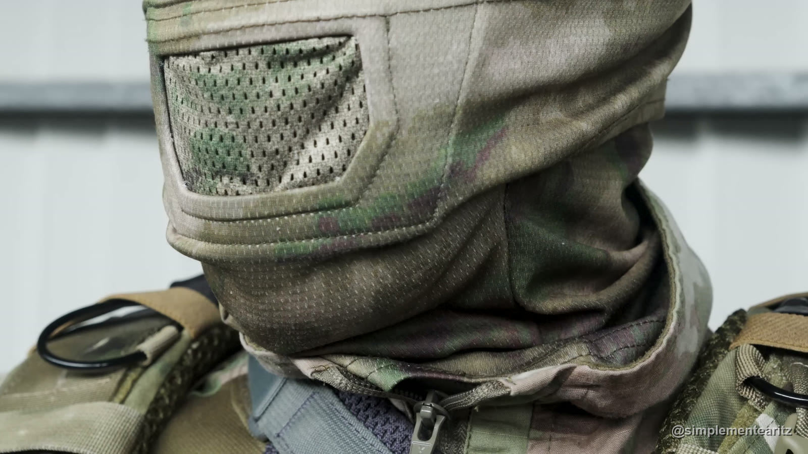 Máscara de tela de Cygnus Armory en camuflaje, puesta, con la rejilla delante de la boca y la tela cubriendo barbilla y cuello, sobre los tirantes de un portaplacas
