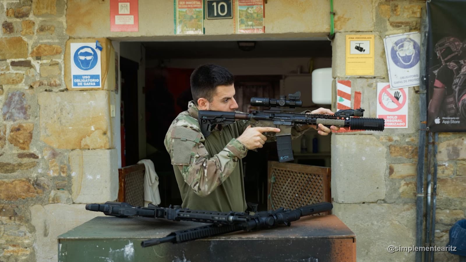 Aritz encarando un fusil de airsoft con mira de aumentos variable, un visor de punto rojo montado encima y un silenciador largo, junto a otras dos réplicas sobre una mesa