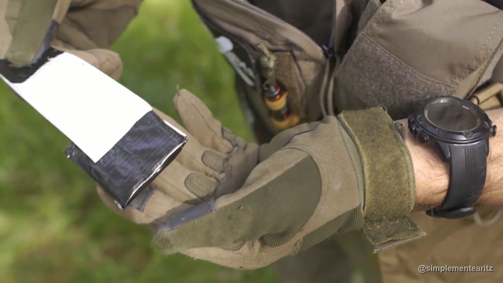 Manos con guantes de airsoft sujetando una tarjeta a medio envolver en cinta americana negra, delante de un chest rig verde oliva