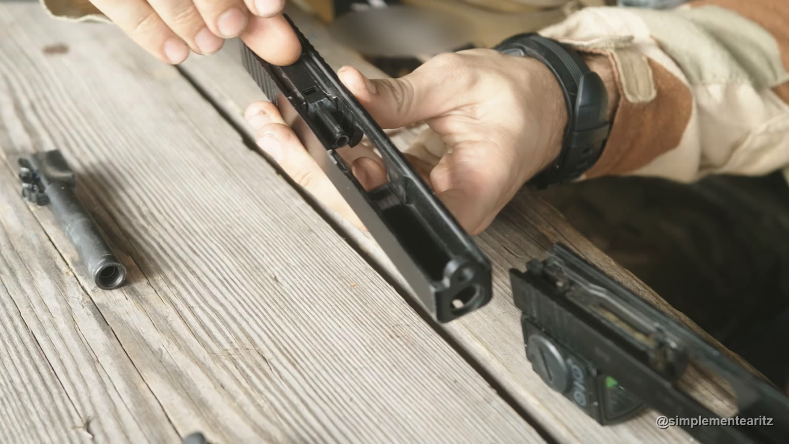 Manos sujetando por debajo la corredera desmontada de una Glock de airsoft de gas, con el nozzle a la vista, sobre una mesa de madera, con el cañón al lado y el armazón con una linterna, desenfocado, en una esquina