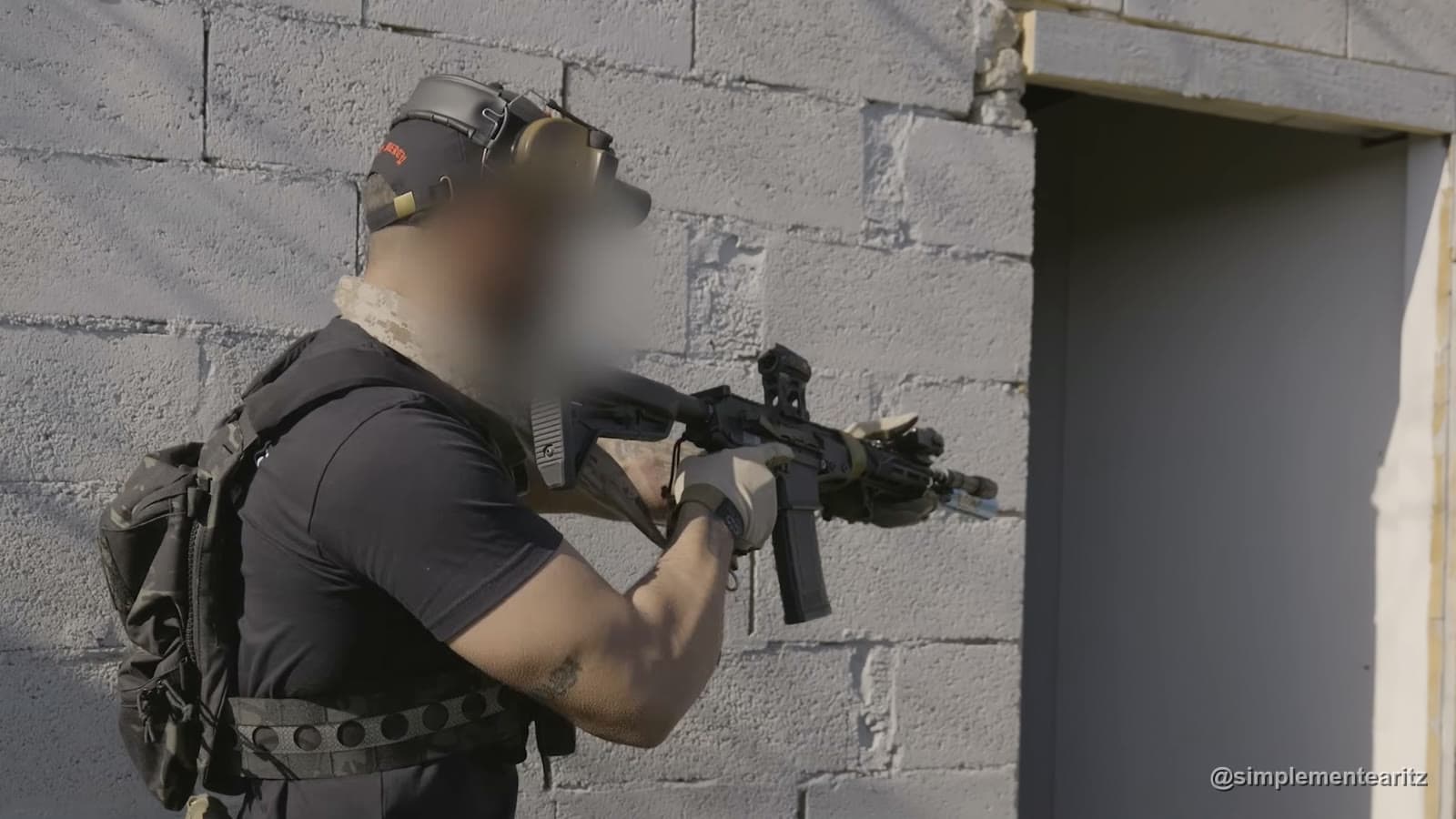 Instructor con gorra, cascos de protección auditiva y mochila acercándose en ángulo a una puerta abierta con un fusil corto encarado, despejando la habitación desde fuera