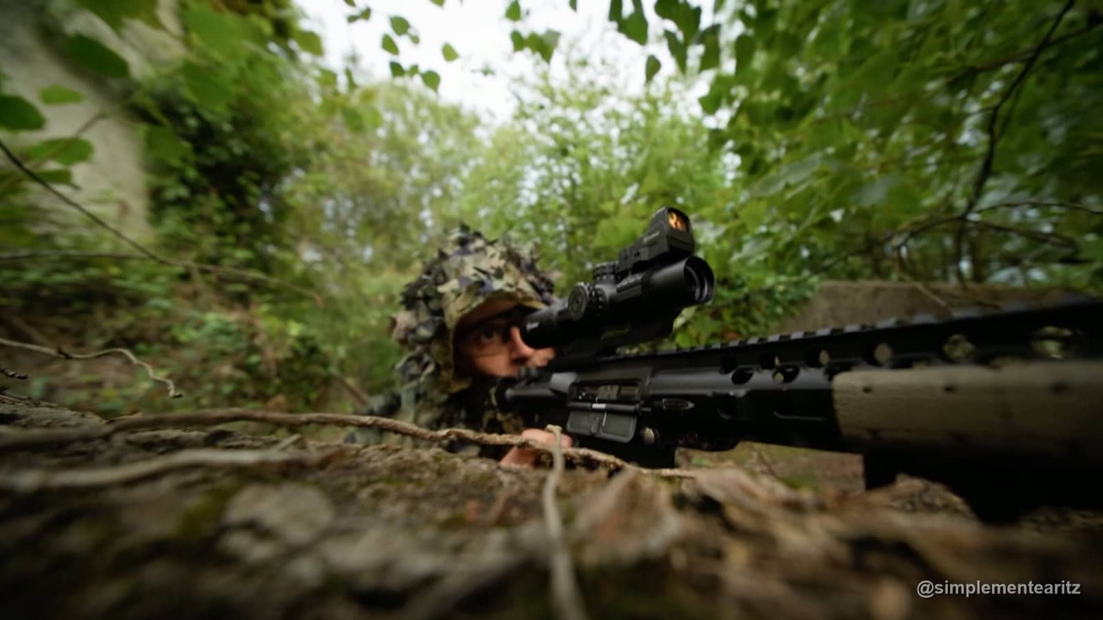 Tirador selecto de airsoft con capucha de camuflaje de hojas, tumbado entre la vegetación y apuntando con un fusil DMR con mira de aumentos y visor de punto rojo