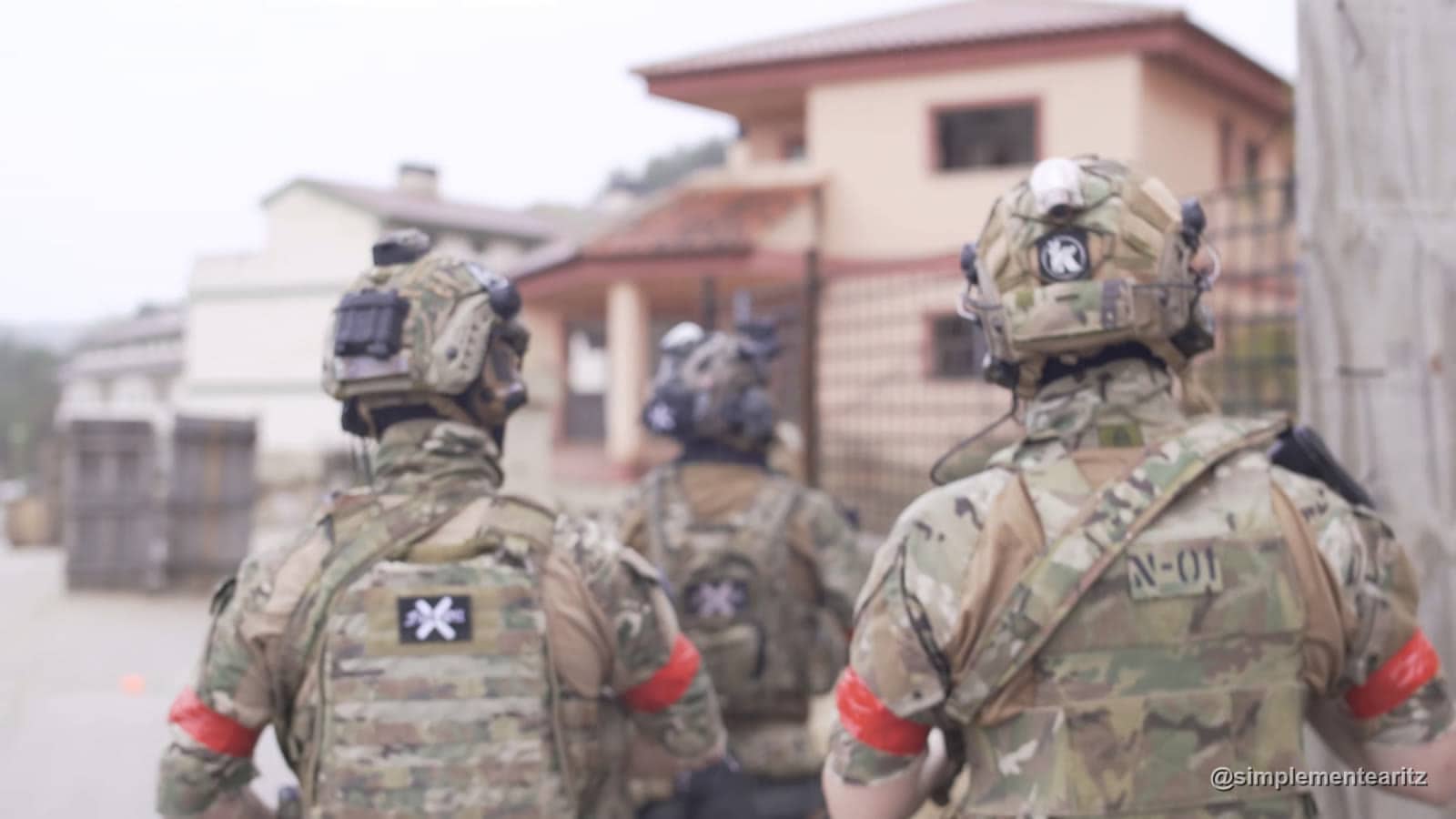 Tres jugadores de airsoft de espaldas con placa portadora multicam y brazalete rojo caminando hacia las casas de una urbanización real en Distrito Zero durante una milsim de tres días