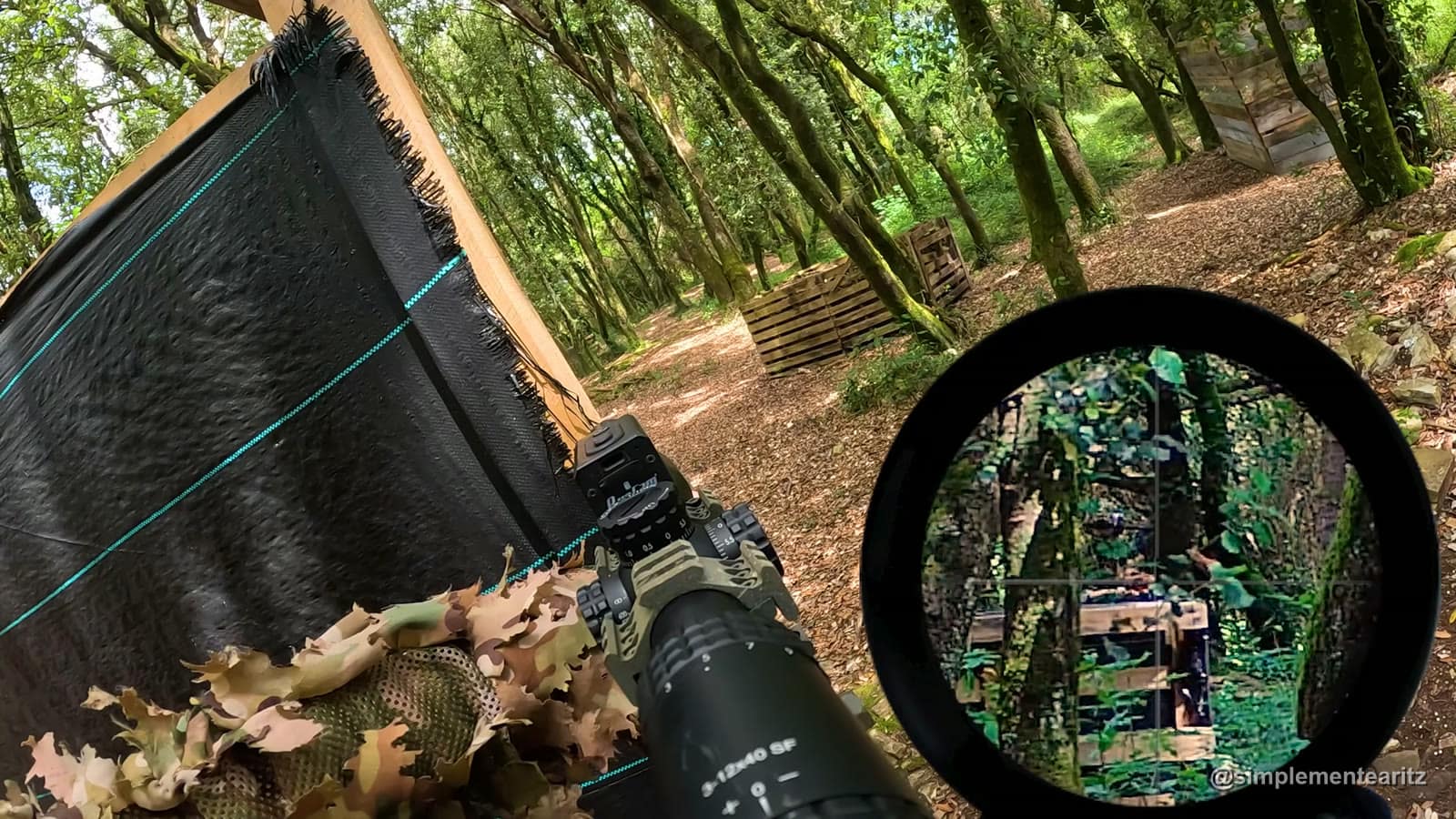 Vista desde detrás de un DMR de airsoft con mira 3-12x40 y punto rojo desplazado, apuntando a unos parapetos de palés en un campo de bosque, con la imagen ampliada de la mira a la derecha