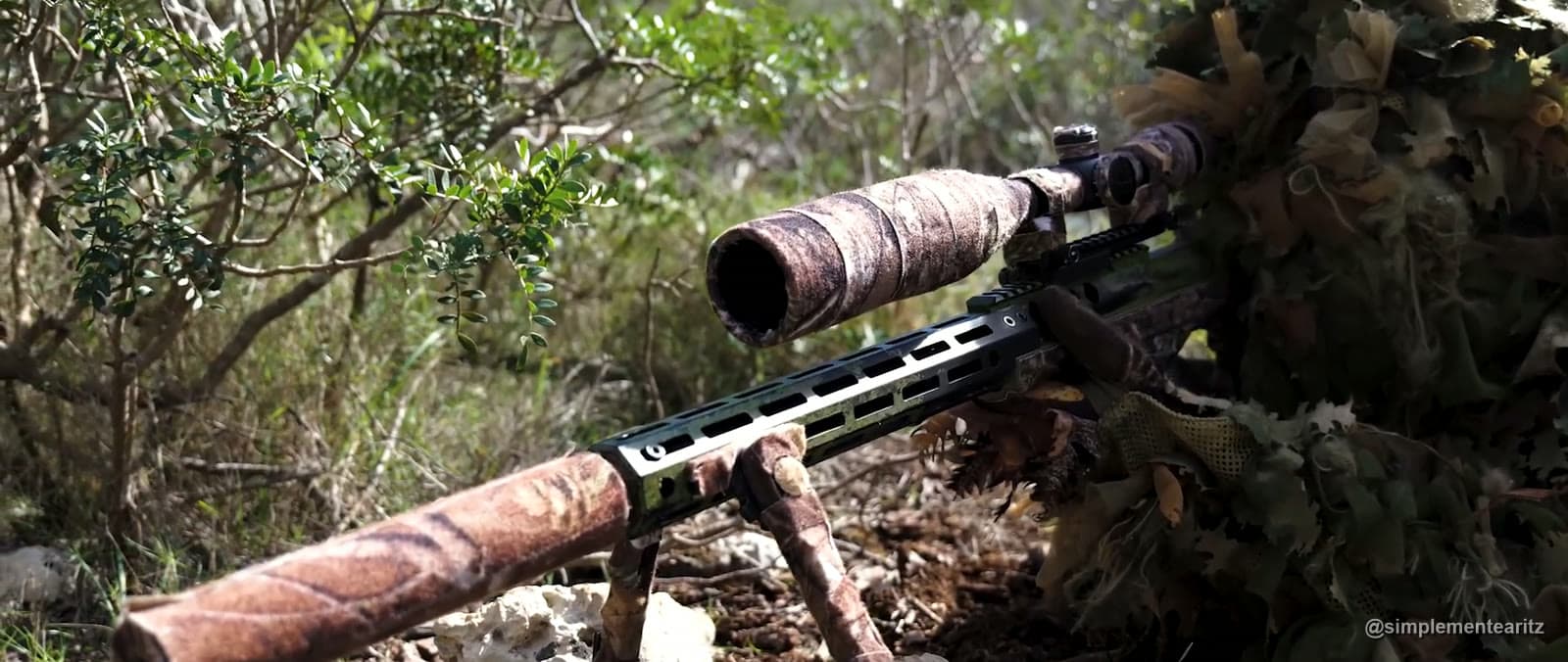 Rifle de francotirador de airsoft con bípode, guardamanos de aluminio y silenciador forrado con cinta de camuflaje, apoyado entre matorrales junto a un tirador con traje ghillie
