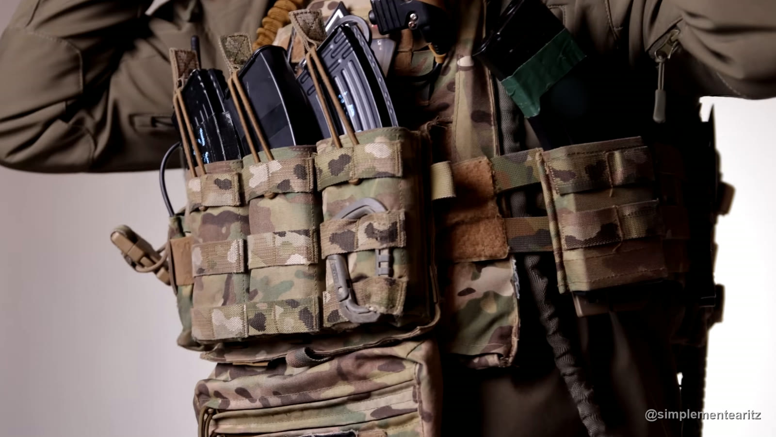 Frontal de un portaplacas puesto sobre una chaqueta marrón verdosa: tres portacargadores abiertos de Warrior Assault Systems en MultiCam con cargadores de AK y un mosquetón, a la derecha el cummerbund del CVS de Conquer, en camuflaje tipo MultiCam, y debajo el bolsillo subabdominal (drop pouch)