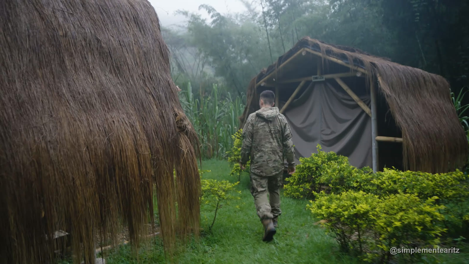 Dos cabañas de bambú con techo de paja, una de ellas cerrada con una lona gris de plástico, en mitad de la selva colombiana con niebla, y un jugador de airsoft con chaqueta de camuflaje caminando de espaldas entre ellas: el «hotel» donde iba a quedar guardado el material