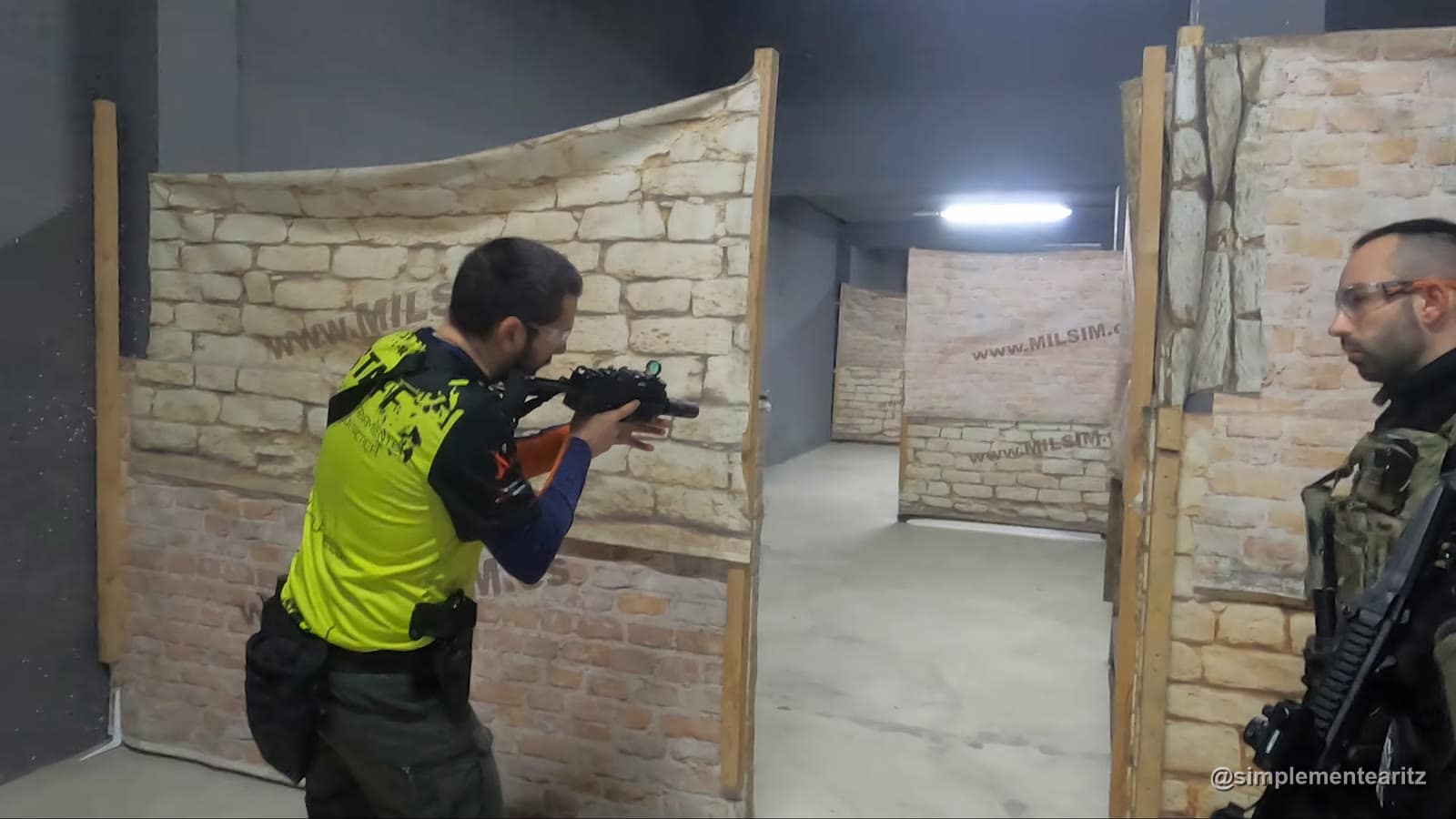 Instructor de El Enclave con camiseta amarilla y un subfusil con visor de punto rojo, pegado al borde de un tabique y apuntando por la esquina de un pasillo de interior, mientras Aritz observa con su réplica
