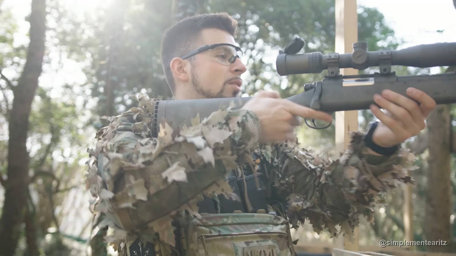 Aritz con chaqueta de camuflaje de hojas sujetando un rifle de francotirador de airsoft de cerrojo con mira telescópica, en un campo entre árboles