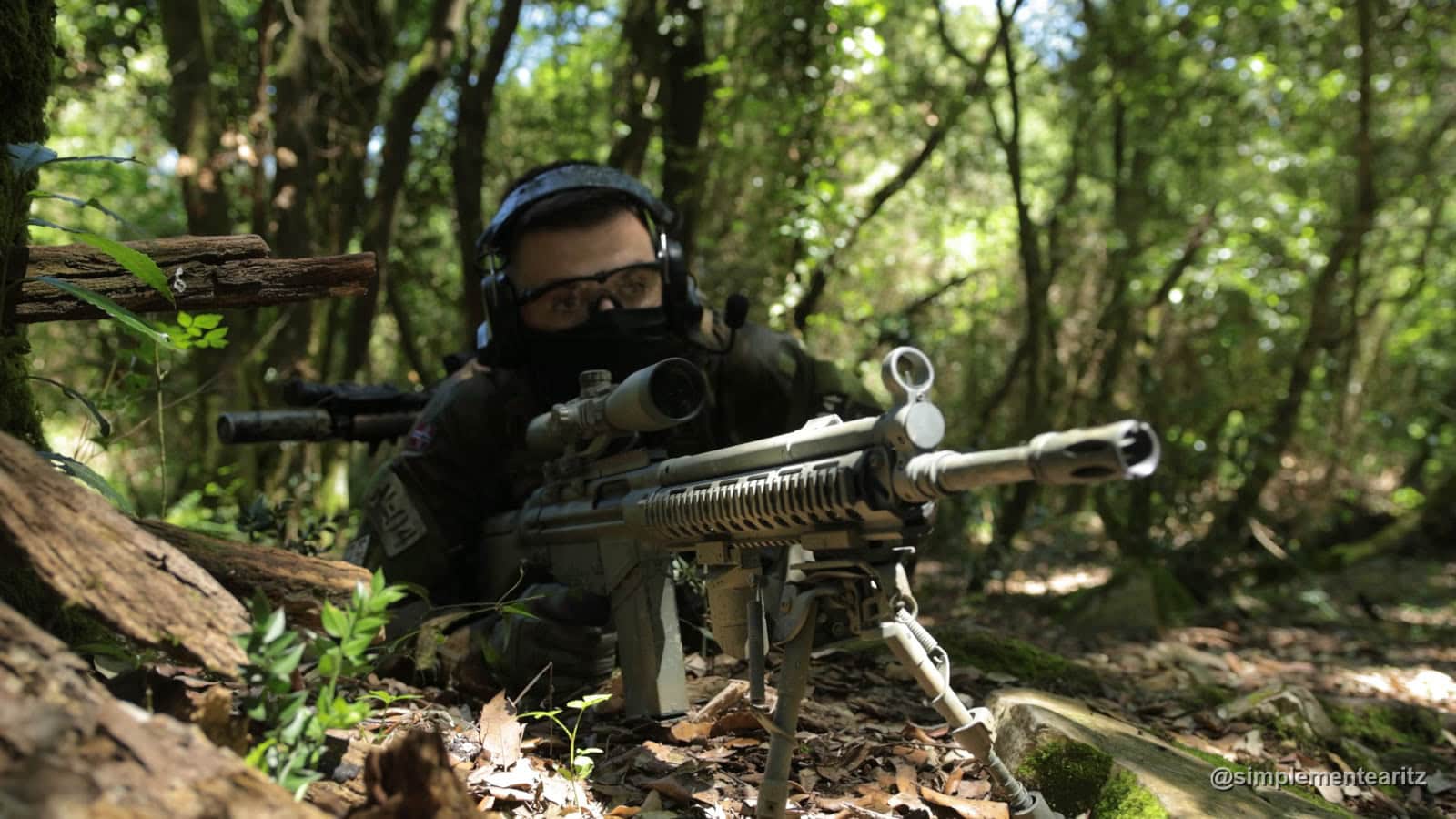 Jugador de airsoft tumbado en el bosque detrás de una réplica tipo G3 de 7,62 con mira telescópica, bípode y guardamanos largo, con un segundo fusil con silenciador a la espalda