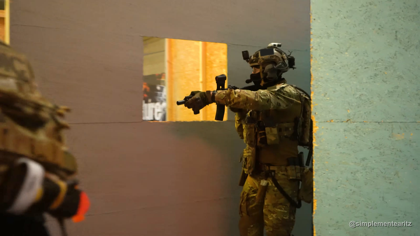 Jugador de airsoft con casco y la cara tapada apuntando una pistola junto al hueco de una ventana, en un CQB de Bélgica