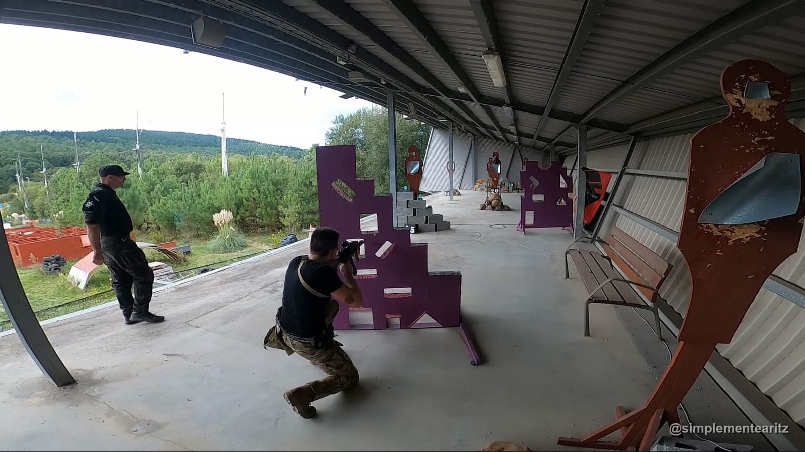 Aritz agachado disparando con un fusil a través de los huecos de un muro de madera morado, en la prueba del muro vikingo de la Copa de Airsoft de Euskadi, con un juez detrás