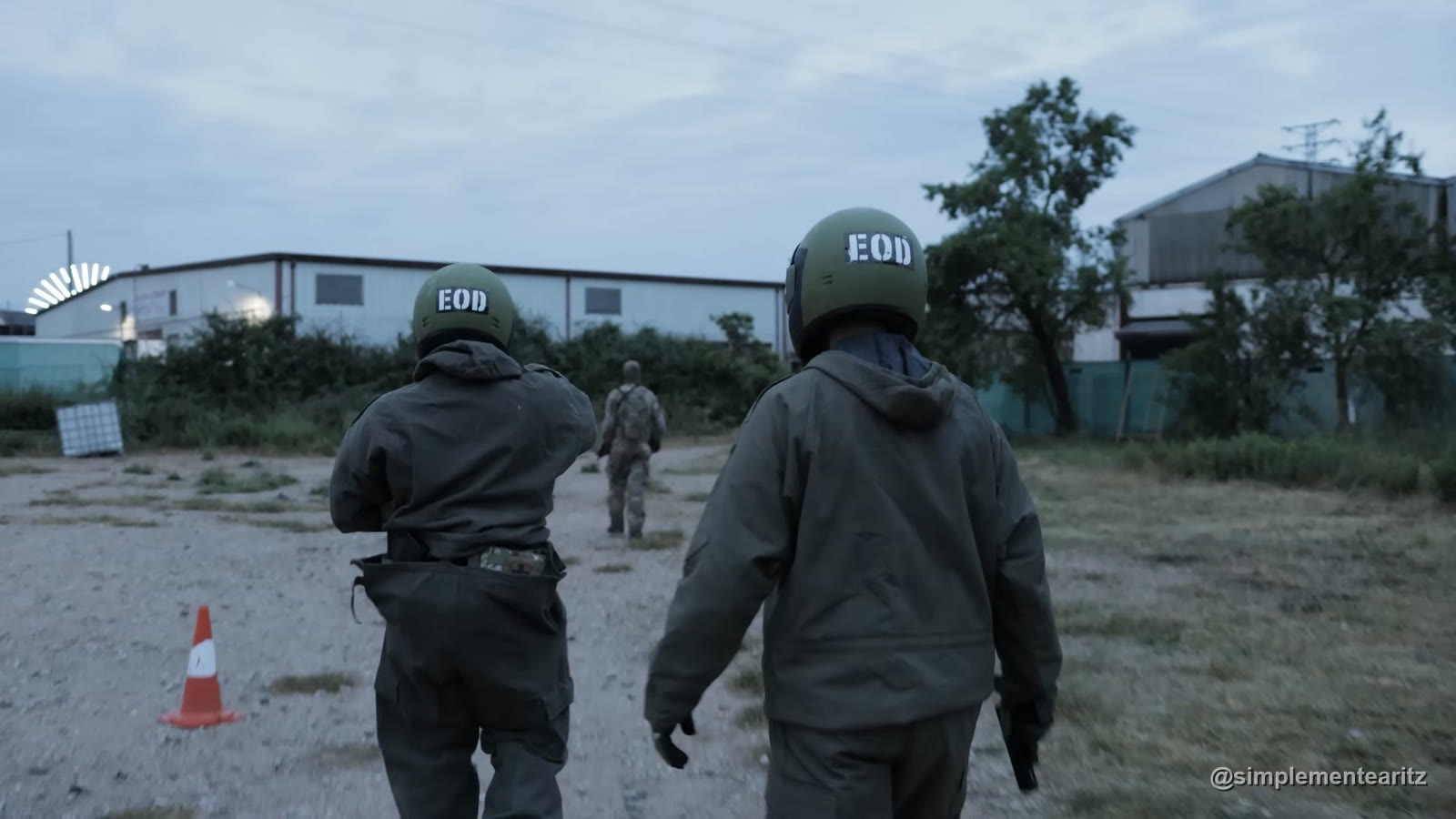 Dos jugadores de airsoft de espaldas con casco verde rotulado EOD y chaqueta verde oliva caminando al atardecer por una explanada de tierra hacia las naves de La Fundición