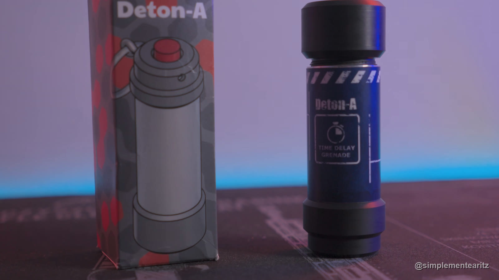 Granada de fogueo Deton-A de pie, negra, con una etiqueta azul en la que pone «Deton-A» y «Time delay grenade», junto a su caja de cartón, sobre una alfombrilla