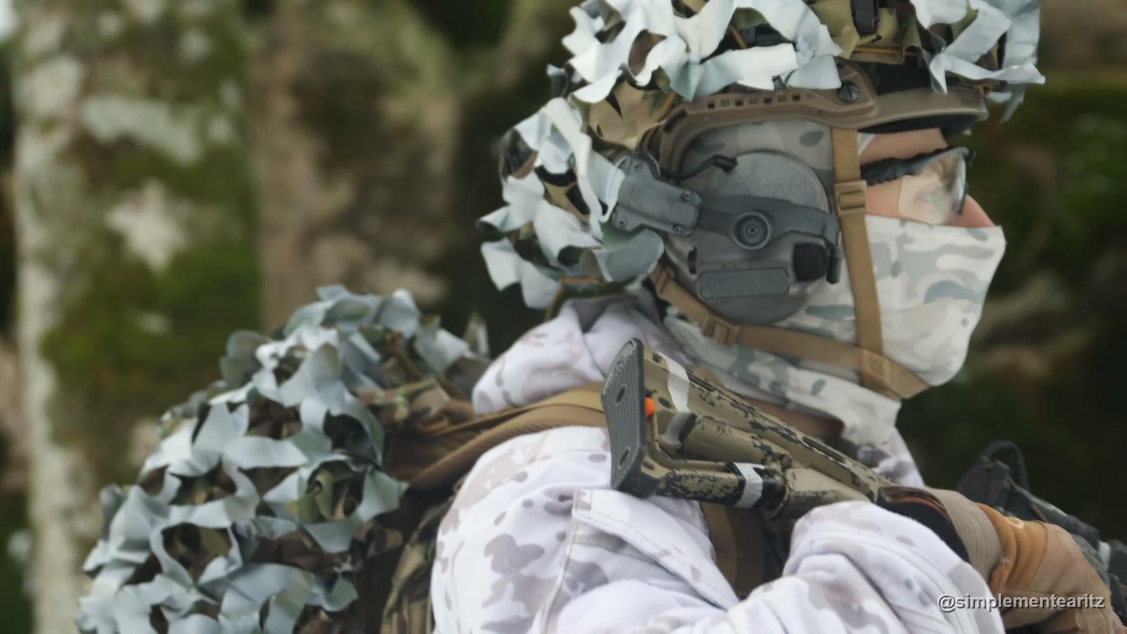 Jugador de airsoft de perfil con un cubrecasco de tiras blancas y de camuflaje, auriculares de comunicación y braga, con la patilla de sus gafas balísticas transparentes perdiéndose bajo la correa del casco, junto al auricular