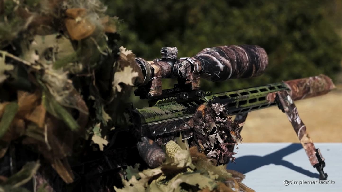 Rifle de francotirador de airsoft con bípode y mira forrados de camuflaje sobre una mesa, junto a la manga de un traje ghillie de hojas