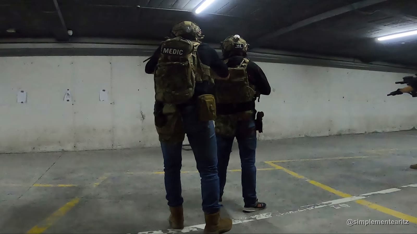 Dos alumnos de espaldas con casco y placa portadora en un entrenamiento de CQB en un aparcamiento, el de atrás pegado al de delante con la mano en su hombro mientras avanzan