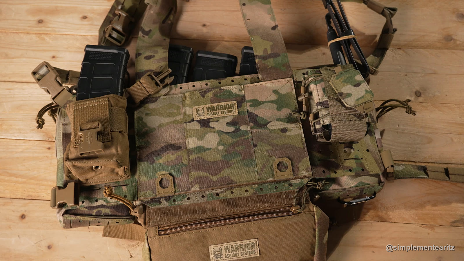 Chest rig MCR MK1 de Warrior Assault Systems en MultiCam sobre una mesa de madera: cargadores de fusil asomando por encima del panel frontal, un bolsillo a cada lado y debajo el bolsillo subabdominal (drop pouch) en coyote. El panel frontal y el bolsillo subabdominal llevan la etiqueta de Warrior