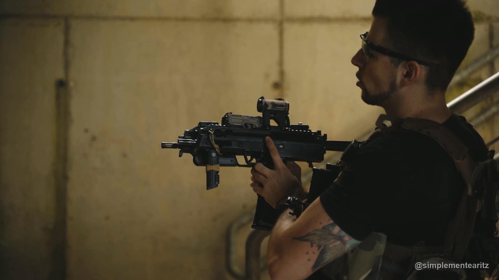 Aritz de perfil, con placa portadora y gafas, apuntando con una MP7 de gas con punto rojo junto a una escalera de hormigón
