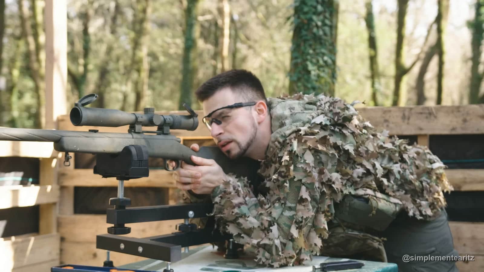 Aritz con chaqueta de camuflaje de hojas apuntando un rifle de francotirador de airsoft tipo M700 con mira telescópica, apoyado en un soporte de tiro sobre una mesa en Max Airsoft