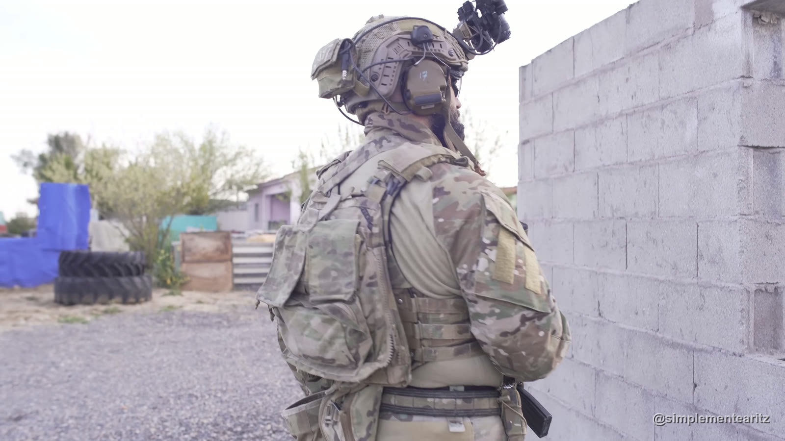 Recreador de airsoft del 75.º Regimiento Ranger de perfil, con uniforme y placa portadora multicam, casco con visor nocturno y velcros del hombro sin parches, junto a un muro de bloques