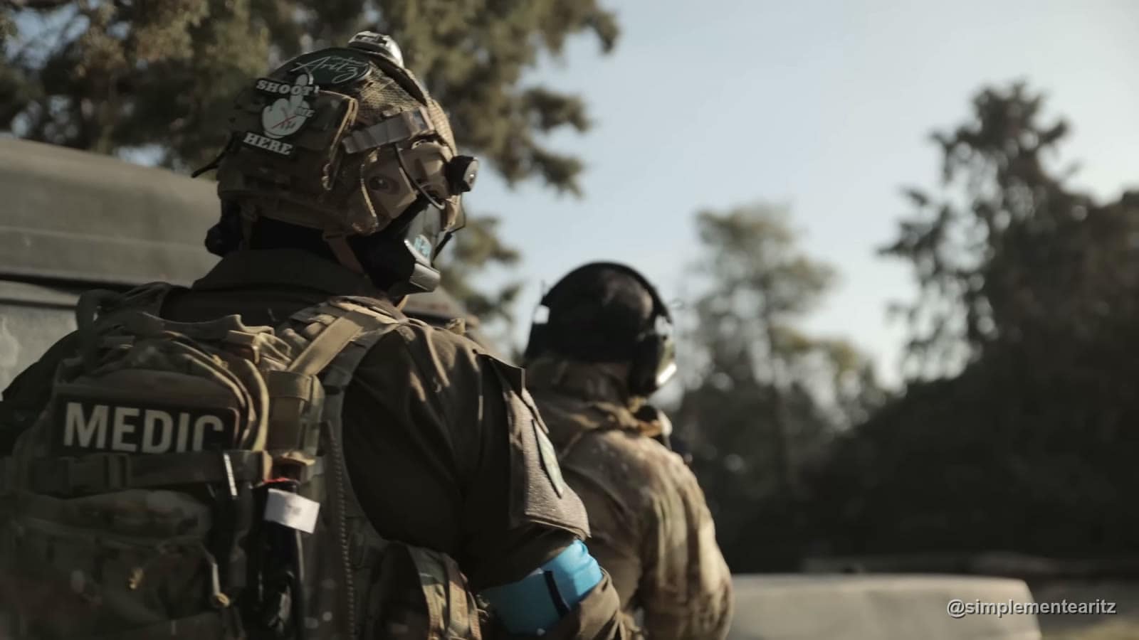 Jugador de airsoft de espaldas con el parche MEDIC en la mochila y un brazalete azul de bando, junto a un compañero con cascos de comunicación